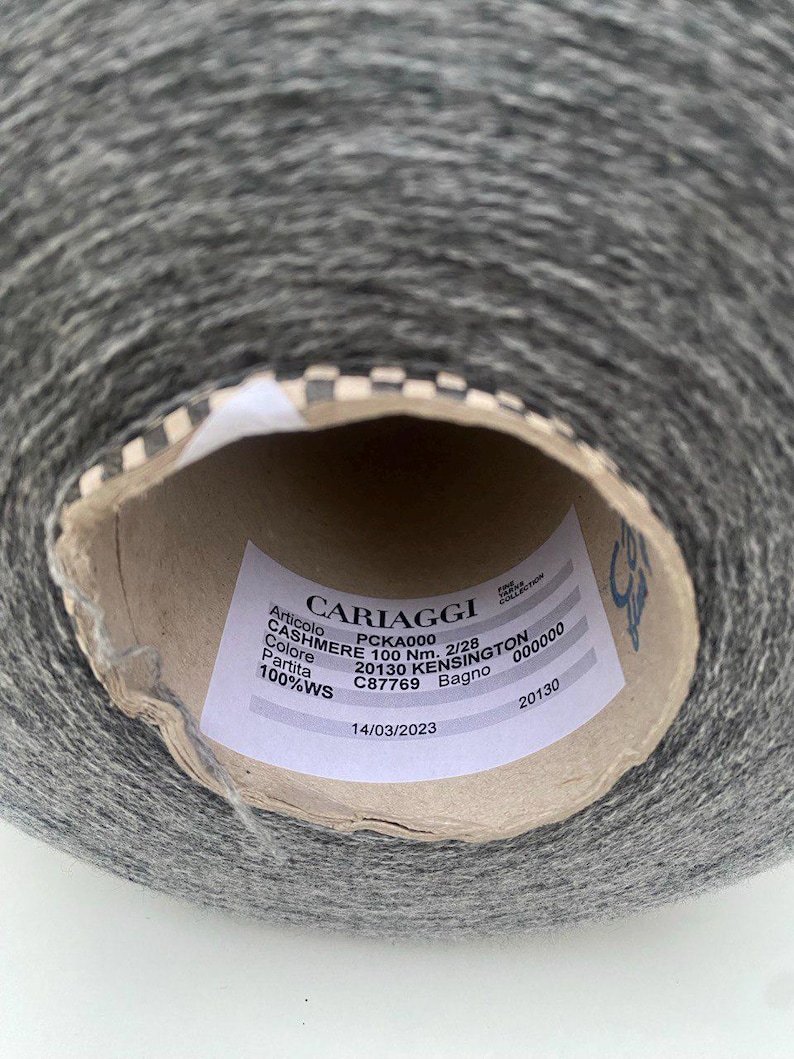 May include: A close-up of a gray yarn ball with a white label that reads "CARIAGGI" and includes details like "PCKA000", "CASHMERE 100 Nm. 2/28", "20130 KENSINGTON", "C87769", "100%WS", and "14/03/2023".