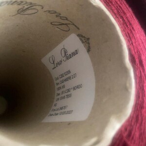 Op de afbeelding: Een close-up van een label op een spoel rood garen. Het label vermeldt "Loro Piana" en bevat details zoals de garencode, de naam, het materiaal, de kleur en de datum.