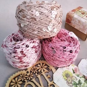 May include: Three skeins of yarn with sequins, one beige, one pink and white, and one pink. The yarn is arranged on a gray surface with a wooden heart that says "Love" and some decorative paper tags.