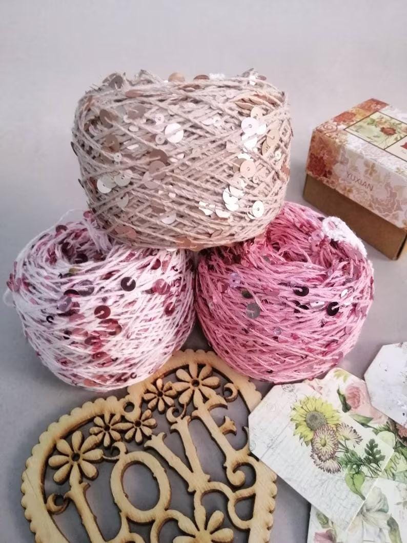 May include: Three skeins of yarn with sequins, one beige, one pink and white, and one pink. The yarn is arranged on a gray surface with a wooden heart that says "Love" and some decorative paper tags.