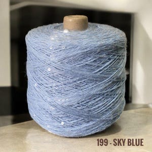 May include: A skein of light blue yarn with white sequins woven throughout. The yarn is wound around a cardboard spool. The label reads "199 - SKY BLUE".