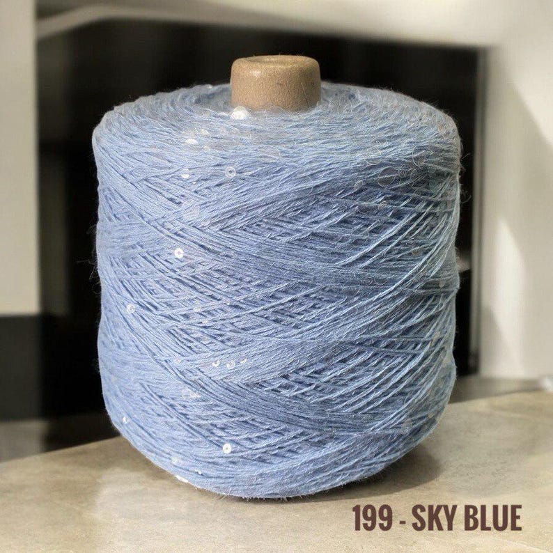 May include: A skein of light blue yarn with white sequins woven throughout. The yarn is wound around a cardboard spool. The label reads "199 - SKY BLUE".
