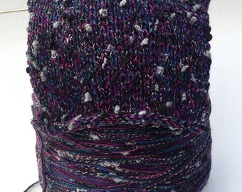 May include: A skein of yarn in a black, purple, blue, and gray colorway with small white and gray flecks throughout. The yarn is wound into a ball and is ready to be used for knitting or crocheting.