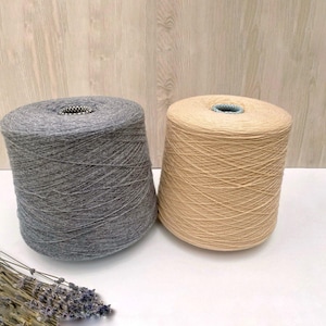 May include: Two spools of yarn, one gray and one beige, are shown on a wooden surface.