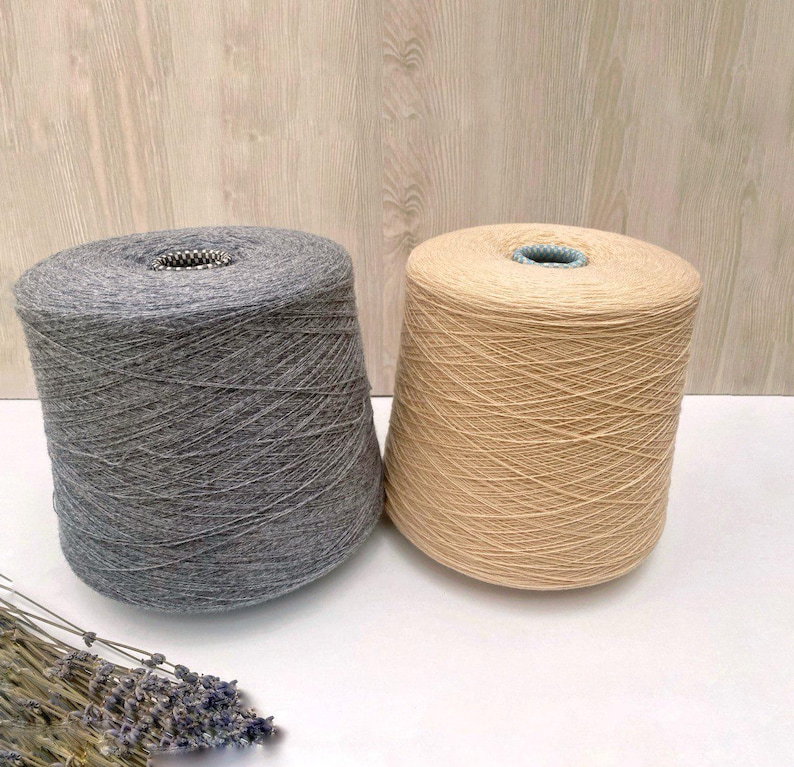 May include: Two spools of yarn, one gray and one beige, are shown on a wooden surface.