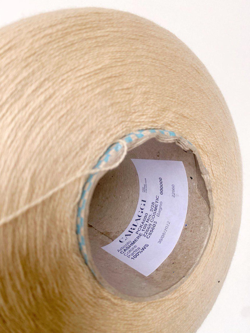 May include: A beige ball of yarn with a blue and white striped label. The label reads "CARIAGGI CASHMERE 100 Nm. 2/28 100%WS Partita PCKA000 22660 COSMETIC C85603 Bagno 000000 22660 30/08/2022".
