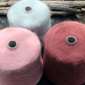 Op de afbeelding: Drie bollen garen in verschillende kleuren: lichtblauw, roze en roodbruin. Het garen is zacht en pluizig en is opgerold in bollen.