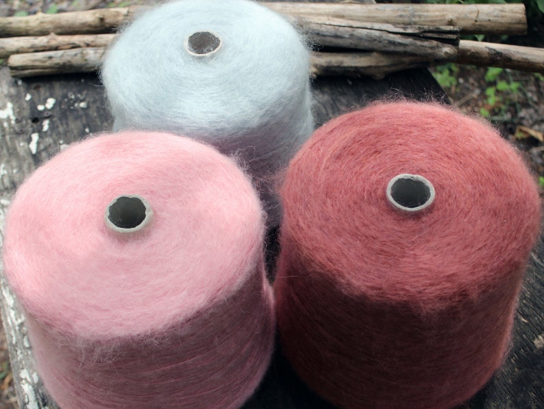 Op de afbeelding: Drie bollen garen in verschillende kleuren: lichtblauw, roze en roodbruin. Het garen is zacht en pluizig en is opgerold in bollen.
