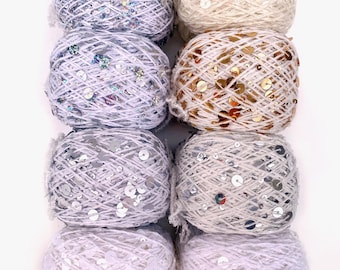 May include: Eight round balls of yarn arranged in two columns, each featuring integrated metallic sequins. The colors include shades of light blue, cream, gold, silver, and white, with each ball showcasing a distinct reflective finish.