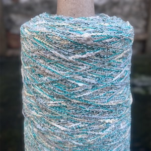 May include: A skein of yarn in a teal, white, and gray color combination. The yarn is textured and fluffy, with a subtle shimmer.