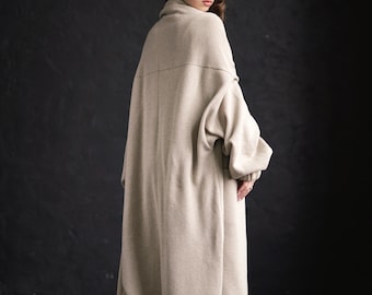May include: A long, beige knitted cardigan with a relaxed fit and a high collar. The cardigan is worn by a person with long brown hair, standing on a dark wooden floor.