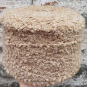 May include: A close-up of a skein of beige fuzzy yarn with a subtle shimmer. The yarn is wound into a tight coil.