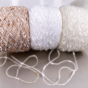 May include: Three balls of yarn in shades of brown, white, and cream. The brown yarn has a speckled appearance. Strands of white yarn are arranged in front of the balls. Suitable for knitting and crochet projects.