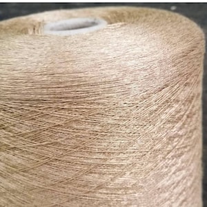 May include: A spool of beige yarn with a subtle shimmer. The yarn is wound tightly and has a smooth texture.