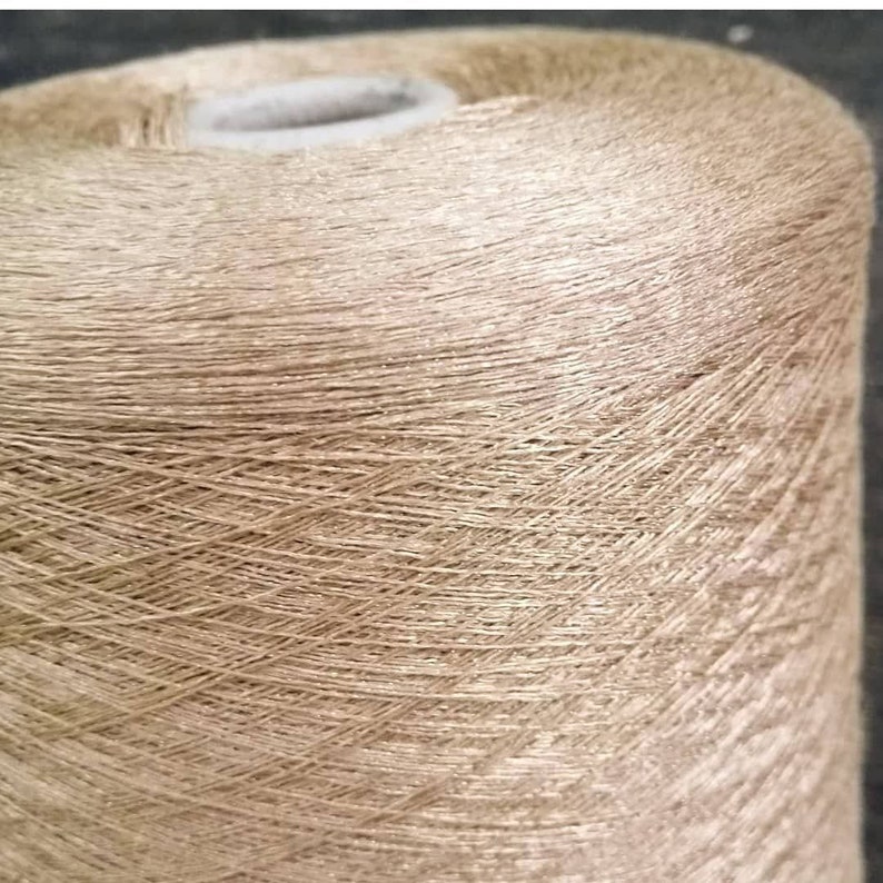May include: A spool of beige yarn with a subtle shimmer. The yarn is wound tightly and has a smooth texture.