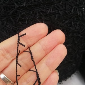 May include: Black yarn with a fuzzy texture and a fringe detail. The yarn is held in a hand.