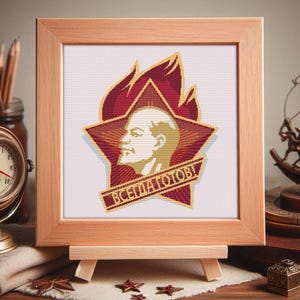 May include: A framed cross-stitch featuring a portrait of a man within a red star and flame design. The text "ВСЕГДА ГОТОВЫ!" is displayed on a banner. The frame is made of light brown wood and is displayed on a wooden easel.