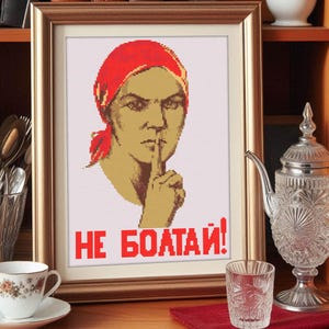 Op de afbeelding: Een ingelijst kruissteekontwerp met een vrouw met een rode hoofddoek en haar vinger op haar lippen. De tekst "НЕ БОЛТАЙ!" staat in rode letters onder de vrouw.