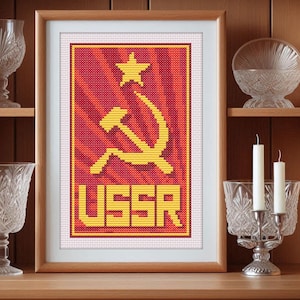 May include: A framed cross-stitch design featuring a red background with a yellow hammer and sickle symbol and a yellow star. The text "USSR" is below the symbol.
