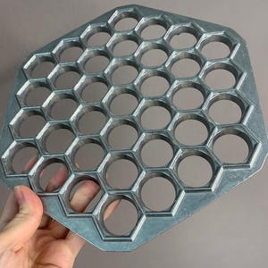 May include: A silver-colored, hexagonal dumpling maker with multiple circular cutouts. The metal kitchen tool is held by a hand, showcasing its honeycomb-like design. This tool is designed for making dumplings.