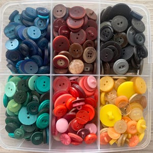 May include: A clear plastic storage container divided into six compartments, each filled with a collection of round buttons in various shades of blue, brown, black, green, red, pink, yellow, and orange, featuring two or four holes.