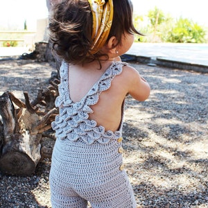 Back view of a toddler wearing a handmade boho crochet romper with scalloped straps and wooden buttons, enjoying an outdoor sunny day. Crochet pattern PDF for babies and toddlers