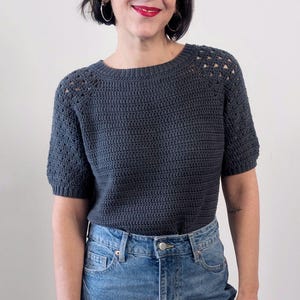 Peut inclure: Un haut en crochet gris foncé à manches courtes et col rond. Le haut présente un motif texturé et des détails ajourés sur les épaules. Il est associé à un jean en denim bleu clair. Le style général est décontracté et confortable.