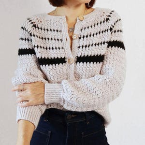 Puede incluir: Un cárdigan de crochet beige claro con rayas negras horizontales en las mangas y el pecho. El cárdigan tiene una parte delantera abotonada y se combina con vaqueros de mezclilla azul oscuro. Las mangas son largas y el cárdigan tiene un patrón texturizado.