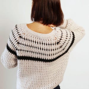 May include: A beige crochet sweater with a black horizontal stripe across the upper back and sleeves. The sweater has a round neckline and a textured stitch pattern. The sleeves are three-quarter length.