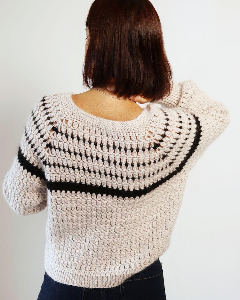 May include: A beige crochet sweater with a black horizontal stripe across the upper back and sleeves. The sweater has a round neckline and a textured stitch pattern. The sleeves are three-quarter length.