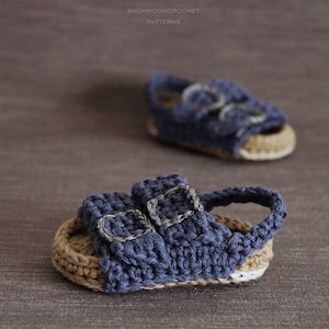 Op de afbeelding: Een paar blauwe gehaakte babysandalen met twee gespen. De sandalen hebben een bruine gehaakte zool.