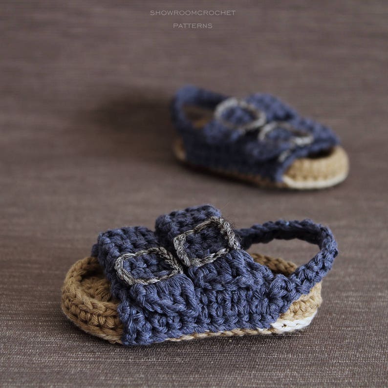 Op de afbeelding: Een paar blauwe gehaakte babysandalen met twee gespen. De sandalen hebben een bruine gehaakte zool.