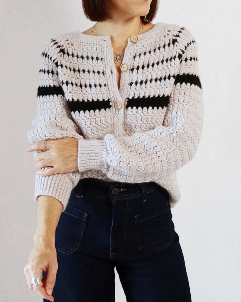 May include: A light beige crochet cardigan with black horizontal stripes on the sleeves and chest. The cardigan has a button-up front and is paired with dark blue denim jeans. The sleeves are long and the cardigan has a textured pattern.