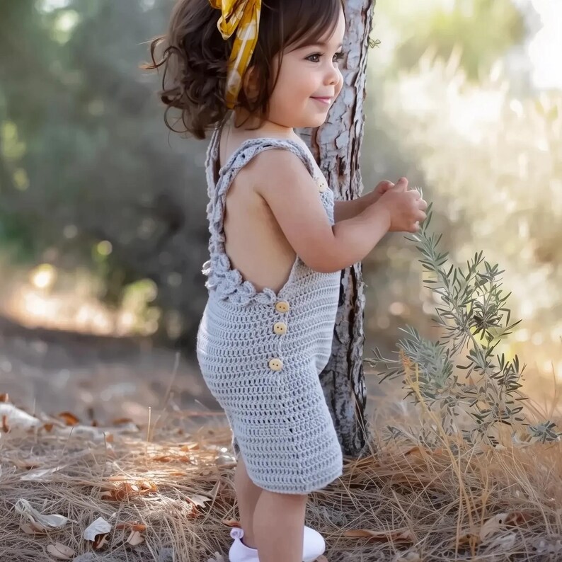 May include: A young child wearing a gray crocheted romper with yellow buttons. The romper has a crocheted flower design on the straps. The child is standing in a grassy area with a tree trunk behind them.