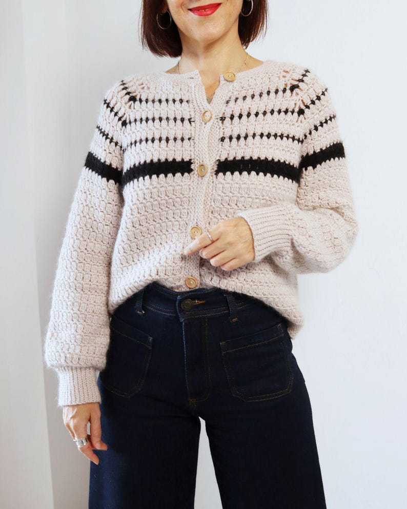 May include: A light beige cardigan with black horizontal stripes and wooden buttons. The cardigan has a textured knit pattern and long sleeves. The person is wearing dark blue jeans.