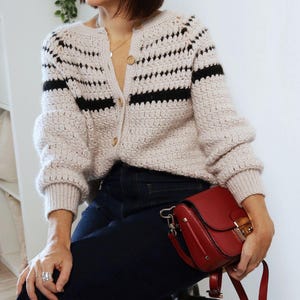 May include: A light beige crochet cardigan with black horizontal stripes and wooden buttons. The cardigan is paired with dark blue jeans and a red leather crossbody bag with a gold clasp.