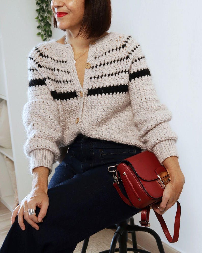 May include: A light beige crochet cardigan with black horizontal stripes and wooden buttons. The cardigan is paired with dark blue jeans and a red leather crossbody bag with a gold clasp.