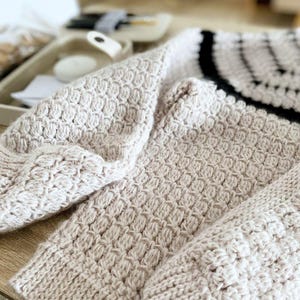 May include: Close-up of a beige knitted sweater with a textured pattern. The sweater features a ribbed cuff and a black and white striped design on the upper back. The soft, warm knitwear is folded on a wooden surface.