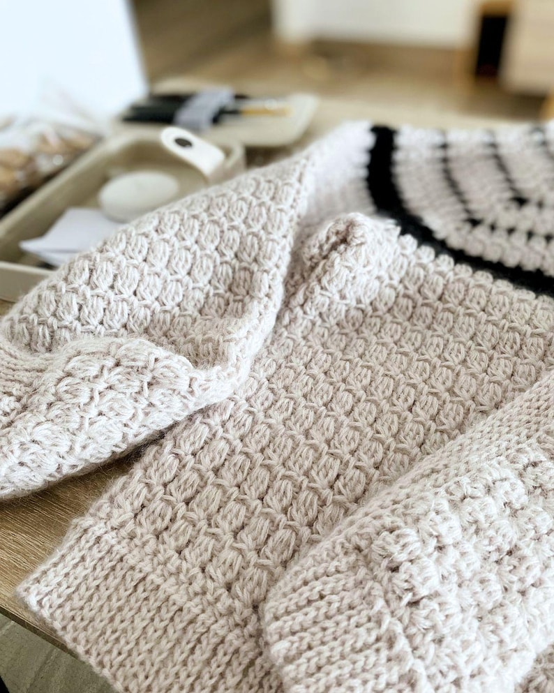 May include: Close-up of a beige knitted sweater with a textured pattern. The sweater features a ribbed cuff and a black and white striped design on the upper back. The soft, warm knitwear is folded on a wooden surface.