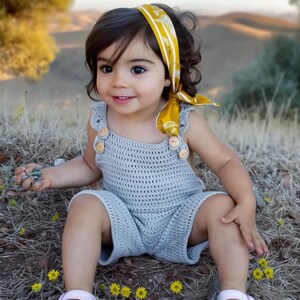 Adorable toddler sitting outdoors wearing a boho crochet romper with wooden buttons. Handmade baby overalls crafted from a detailed crochet pattern PDF, available in sizes 0-8 years