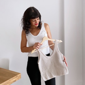 Woman holding a beige crochet tank top on a wooden hanger while wearing a white handmade crochet top made using the Top Cora pattern. This stylish summer crochet pattern is perfect for casual outfits and is crafted with lightweight DK yarn.