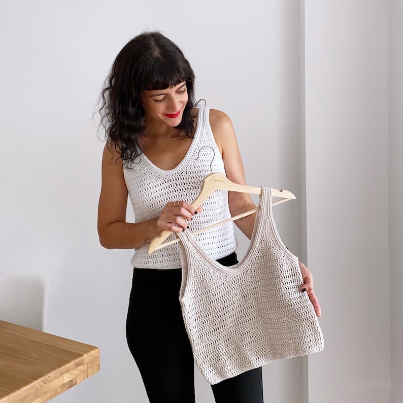 Woman holding a beige crochet tank top on a wooden hanger while wearing a white handmade crochet top made using the Top Cora pattern. This stylish summer crochet pattern is perfect for casual outfits and is crafted with lightweight DK yarn.