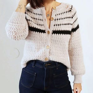 May include: A light beige crochet cardigan with black horizontal stripes and wooden buttons. The cardigan has long sleeves with a textured pattern and is paired with dark blue denim jeans.