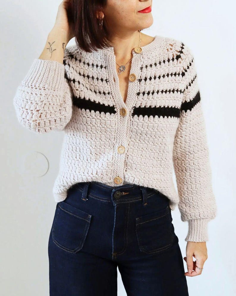May include: A light beige crochet cardigan with black horizontal stripes and wooden buttons. The cardigan has long sleeves with a textured pattern and is paired with dark blue denim jeans.