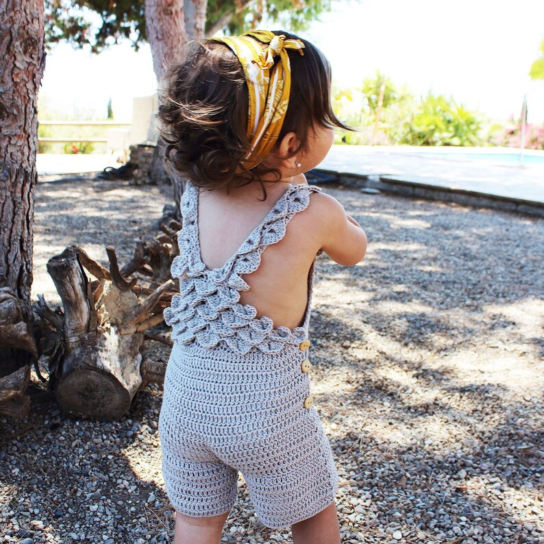 Back view of a toddler wearing a handmade boho crochet romper with scalloped straps and wooden buttons, enjoying an outdoor sunny day. Crochet pattern PDF for babies and toddlers