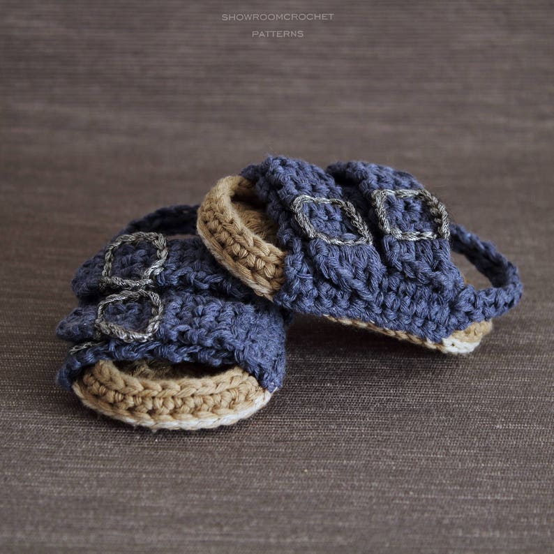 Op de afbeelding: Een paar gehaakte babysandalen in blauw en bruin met zilveren gespen. De sandalen zijn gemaakt met een textuur-steek en hebben een eenvoudig ontwerp.