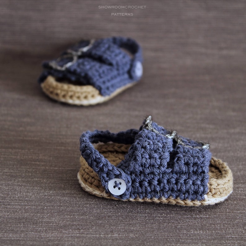 Op de afbeelding: Een paar blauwe gehaakte babysandalen met een bruine gehaakte zool. De sandalen hebben een gespsluiting en een knoopdetail.