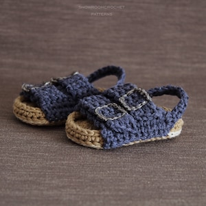 Op de afbeelding: Een paar gehaakte babysandalen met blauwe banden en bruine zolen. De sandalen hebben zilveren gespen.