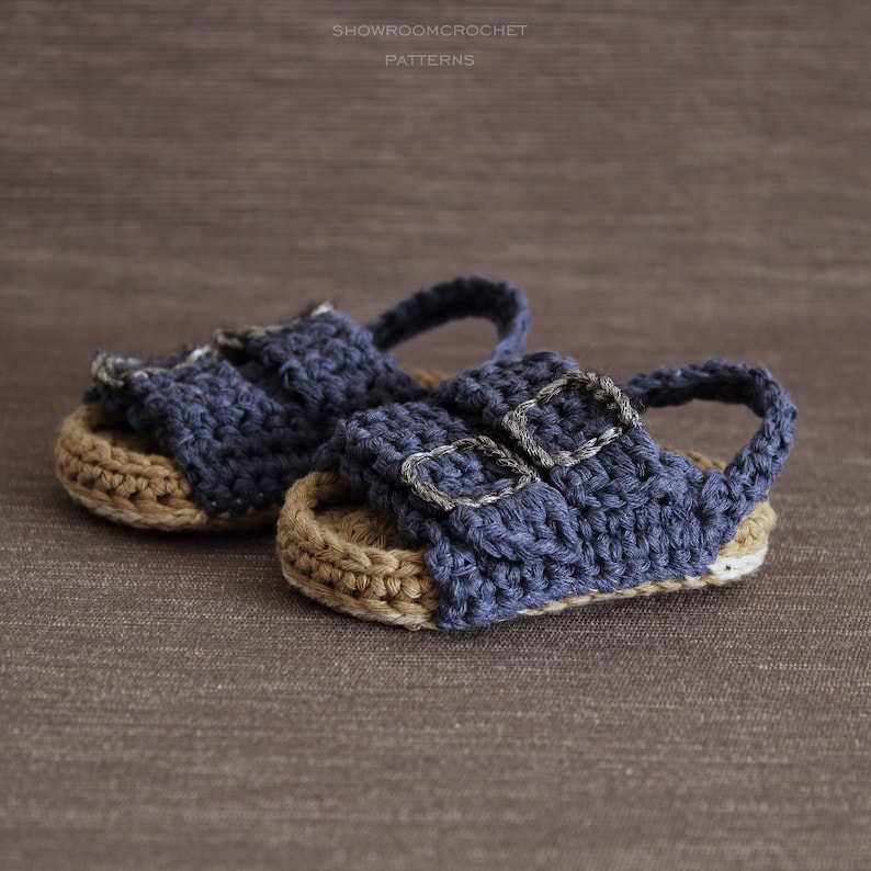 Op de afbeelding: Een paar gehaakte babysandalen met blauwe banden en bruine zolen. De sandalen hebben zilveren gespen.