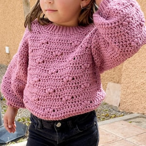 Può includere: Un maglione rosa lavorato a uncinetto con un motivo testurizzato e maniche a sbuffo.